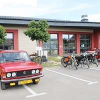 Zdjęcie parkingu przed biblioteką, na którym ustawiono pojazdy z okresu PRL-u, widać czerwonego Fiata 125p zwanego potocznie "dużym fiatem" oraz kilka motocykli.