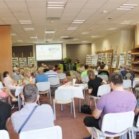 Zdjęcie czytelni biblioteki gdzie odbywało się spotkanie, uczestnicy siedzą przy stolikach i słuchają zaproszonych gości.