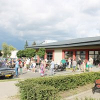 Zdjęcie parkingu przed biblioteką, widać zebranych uczestników spotkania oglądających pojazdy z okresu PRL-u.