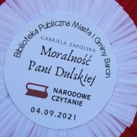 Kotylion przygotowany na Narodowe Czytanie, zawiera napisy Biblioteka Publiczna Miasta i Gminy Barcin ułożony po łuku do góry, Gabriela Zapolska Moralność Pani Dulskiej umieszczony na środku, po niżej tego napisu znajduje się logo akcji, jest to biało-czerwona flaga z dwurzędowym napisem Narodowe Czytanie, na samym dole znajduje się data 04.09.2021r.