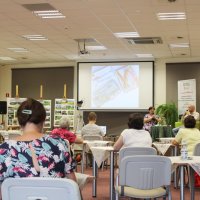 Zdjęcie z tyłu sali, widać uczestników siedzących przy stolikach, stoliki nakryte są serwetkami, na ekranie projektora jest wyświetlany pokaz slajdów złożony ze zdjęć przywiezionych przez Pana Przylipiaka.