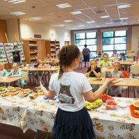 Pani Marzena Żurek stoi za stołami, na których leżą różne owoce i opowiada dzieciom o zdrowym odżywianiu.