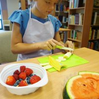 Dziewczynka kroi w plasterki banana, na talerzyku ma położonego arbuza a w miseczce leżą truskawki, borówki i maliny.