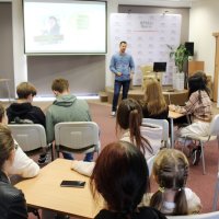 Zdjęcie z głębi sali widać 10 osób siedzących przy stolikach, oraz prowadzącego stojącego przed nimi.