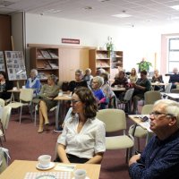 Zdjęcie publiczności, widać dwadzieścia osób, jedna stoi z tyłu sali pozostałe siedzą przy stolikach i słuchają prelegenta.