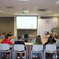 Zdjęcie z tyłu czytelni w bibliotece pokazujące uczniów siedzących przy stołach ustawionych w kształcie podkowy i prowadzącą zajęcia bibliotekarkę siedzącą na krześle przed uczniami.