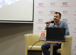 Maciej Piera siedzi w fotelu ustawionym na scenie i mówi do mikrofonu trzymanego w ręku, przed nim na stoliku znajduje się jego książka i laptop.