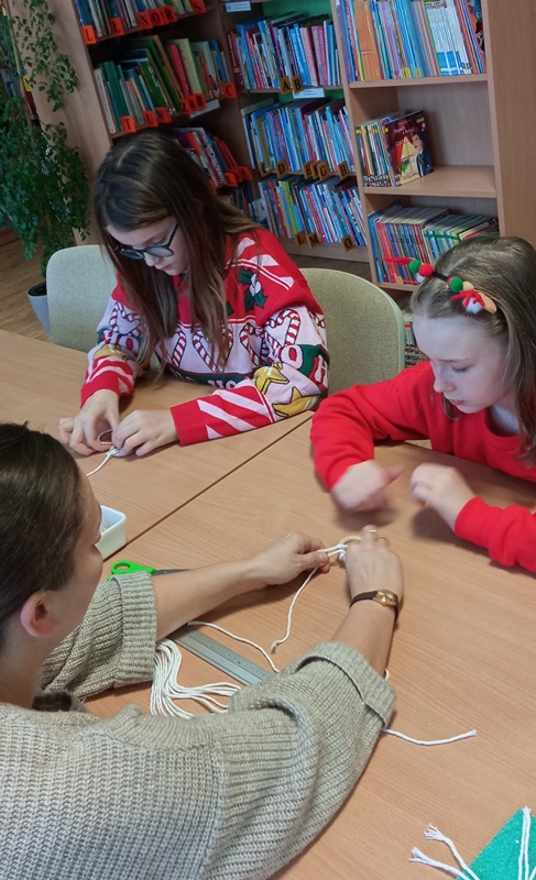 Dwie uczestniczki warsztatów siedzą przy stole razem z bibliotekarką i pracują