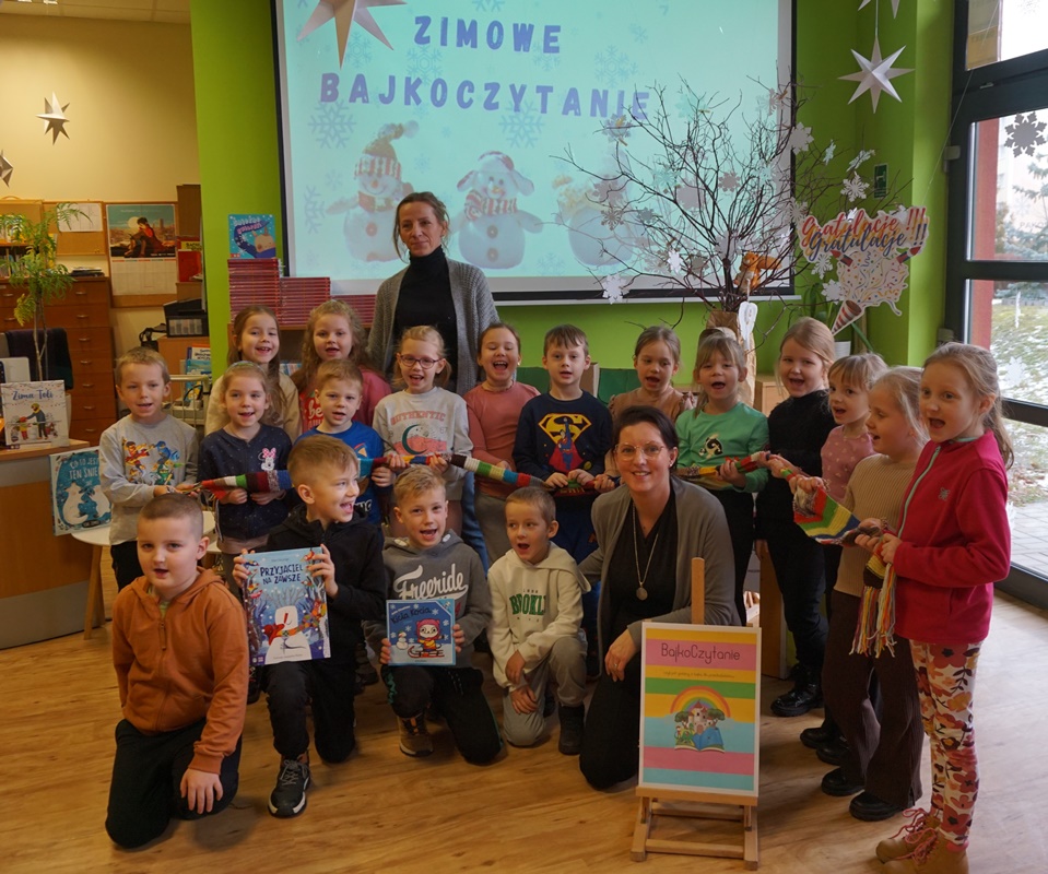 zdjęcie grupy dzieci z opiekunką i bibliotekarką prowadzącą zajęcia, dzieci trzymają książki z przodu stoi kolorowy plakat z napisem bajkoczytanie