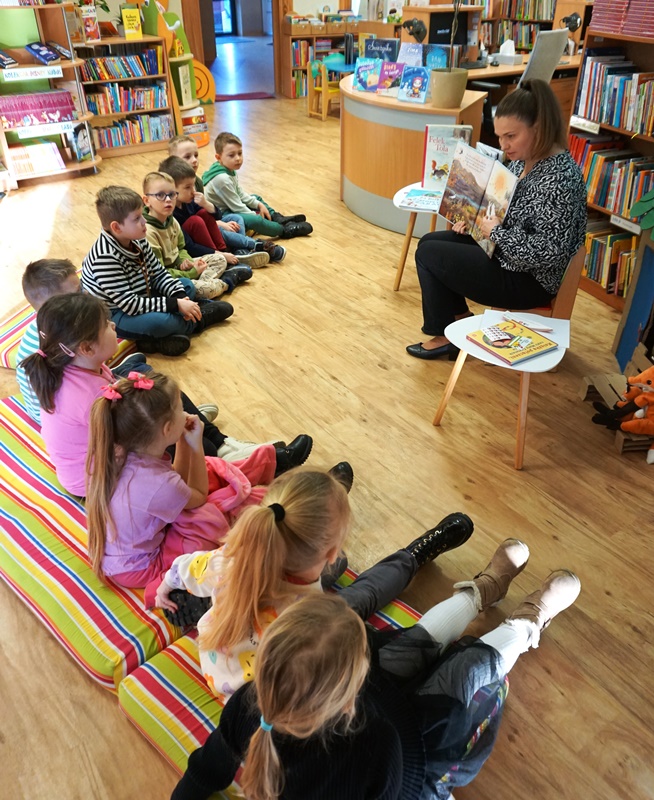 dzieci siedzą na kolorowym materacu, bibliotekarka czyta im książkę