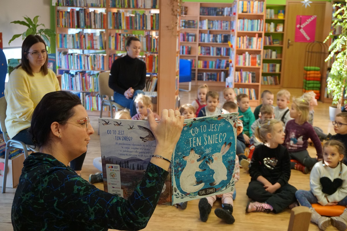 bibliotekarka pokazuje dzieciom wnętrze czytanej książki, na okładce widać białego zająca i lisa