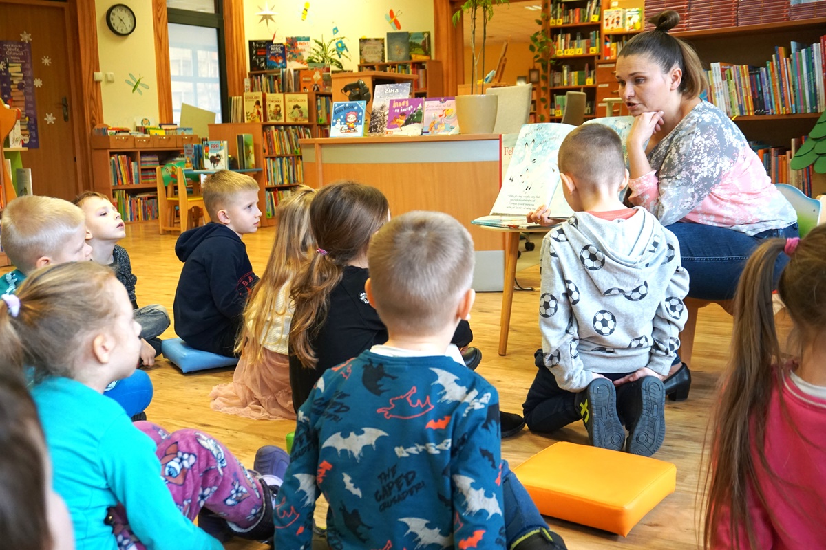 bibliotekarka trzymająca otwartą książkę skierowaną w stronę dzieci które siedzą przed nią i słuchają z uwagą czytanej książki
