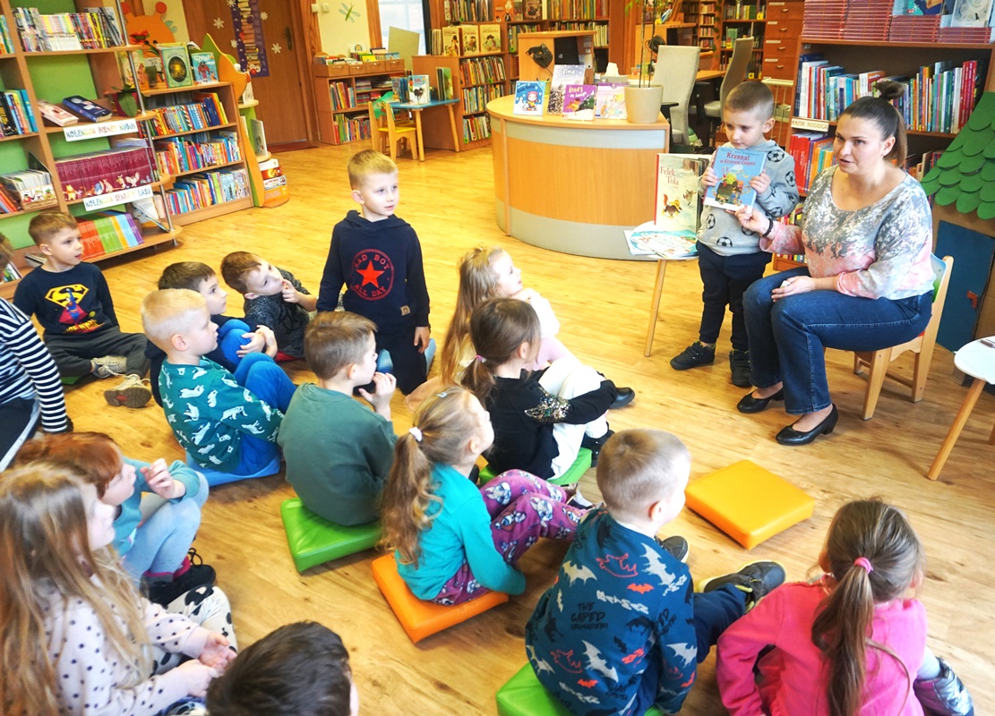 dzieci siedzące na kolorowych pufkach słuchające pani bibliotekarki.