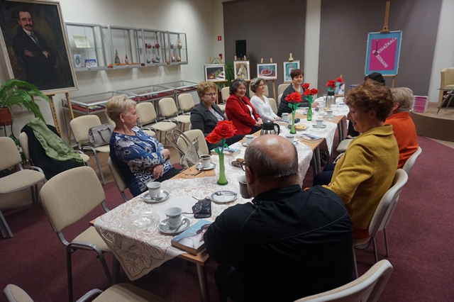 Uczestnicy spotkania siedzą zebrani wokół stołów ustawionych w rzędzie, na stole znajduje się poczęstunek w formie kawy, herbaty i ciastek, stół ozdabiają białe serwetki i wazoniki z papierowymi różami.