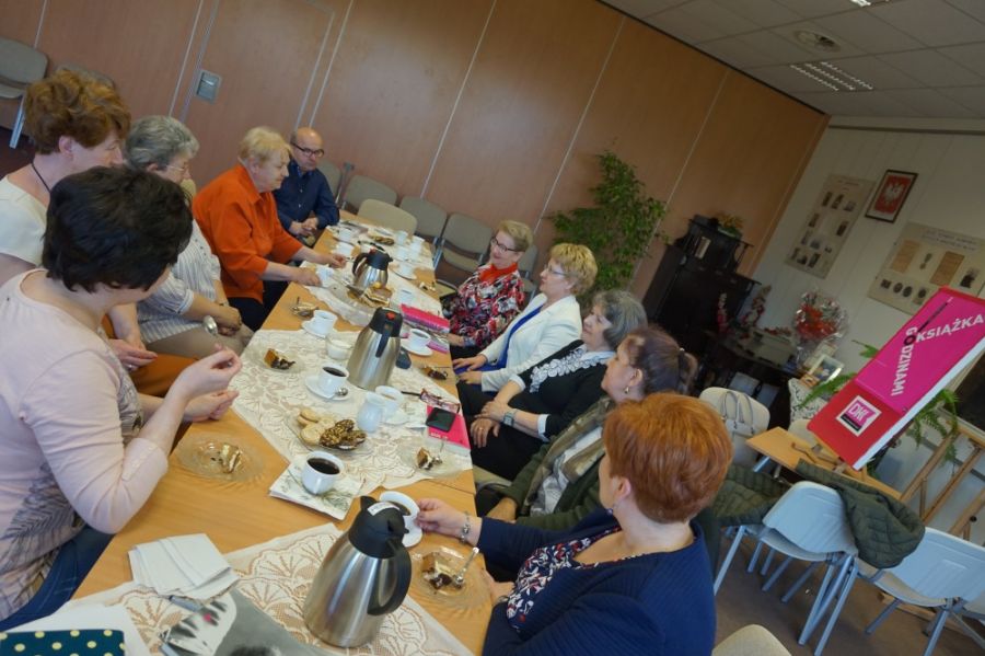 Dziewięć kobiet i jeden mężczyzna obecni na spotkaniu siedzą wzdłuż stołów zastawionych kawą, herbatą i ciastkami, rozmawiają między sobą, po prawej stronie zdjęcia widać plakat DKK stojący na sztaludze