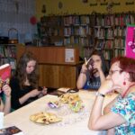 4 czytelniczki i prowadząca spotkanie bibliotekarka siedzą przy stołach i czytają książki, cześć dziewczyn zakrywa twarze otwartymi ksiażkami.