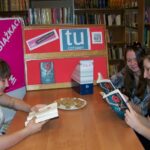 3 nastolatki siedzą przy stole i czytają ksiażki, w tle widać plakat z napisem Godzinami o książkach, tablicę z logo DKK i Instytutu Ksiażki
