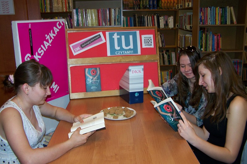 3 nastolatki siedzą przy stole i czytają ksiażki, w tle widać plakat z napisem Godzinami o książkach, tablicę z logo DKK i Instytutu Ksiażki
