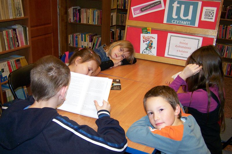 5 dzieci siedzi przy stole, jeden z chłopców czyta książkę, pozostałe dzieci słuchają czytającego chłopca