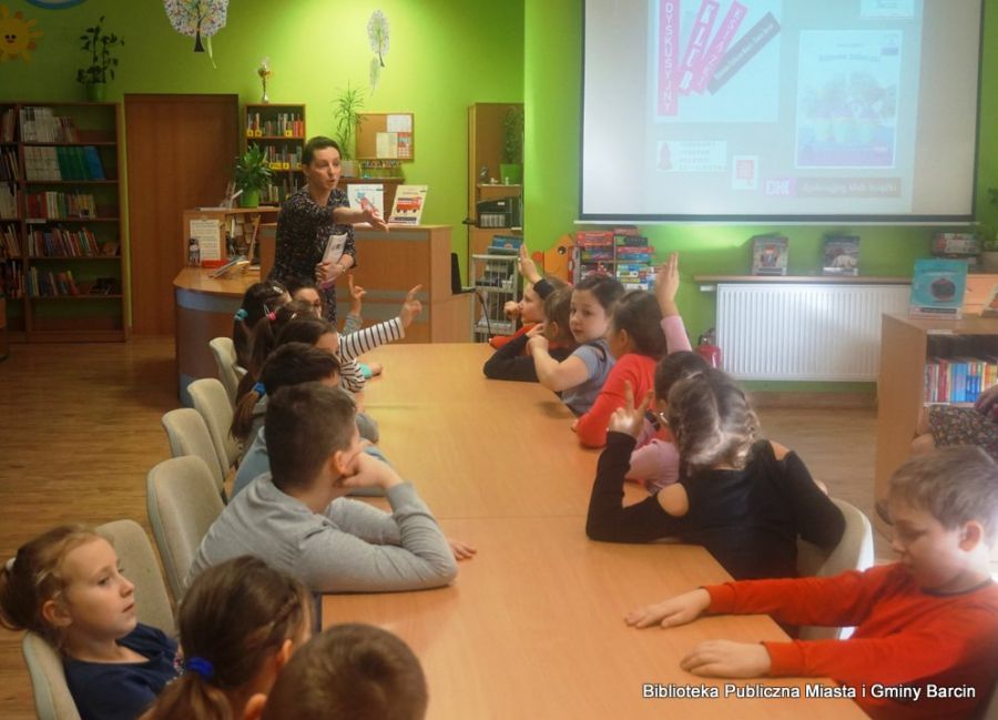 15 dzieci siedzi po dwóch stronach stołów ustawionych w rzędzie, bibliotekarka prowadząca spotkanie wskazuje ręką na jedno z dzieci.