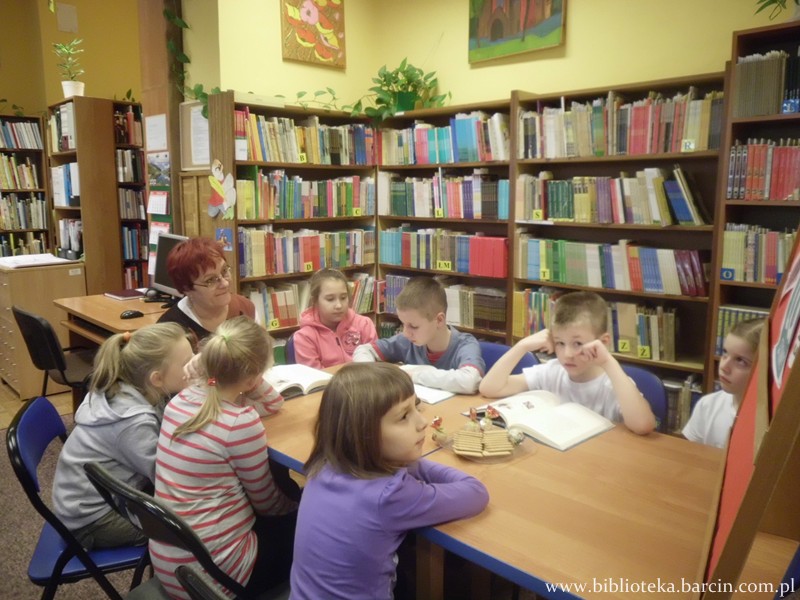 7 dzieci siedzi razem z prowadząca spotkanie bibliotekarką siedzą przy stołach, 2 chłopców i prowadząca mają przed sobą otwarte ksiażki.
