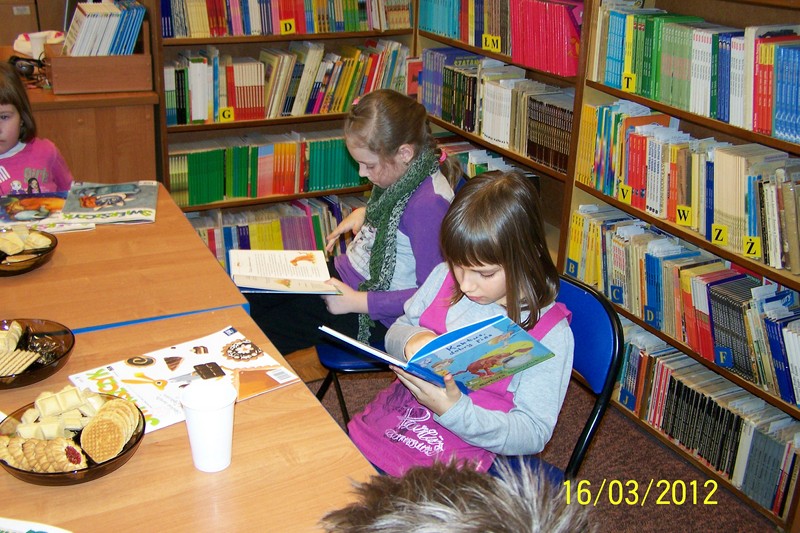 2 dziewczynki siedzą na krzesłach i czytają książki, przed nimi na stołach leżą ciastka, kolorowe czasopisma dla dzieci i jednorazowe kubki