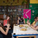 4 czytelniczki i prowadząca spotkanie bibliotekarka siedzą przy stołach i czytają książki, cześć dziewczyn zakrywa twarze otwartymi ksiażkami.