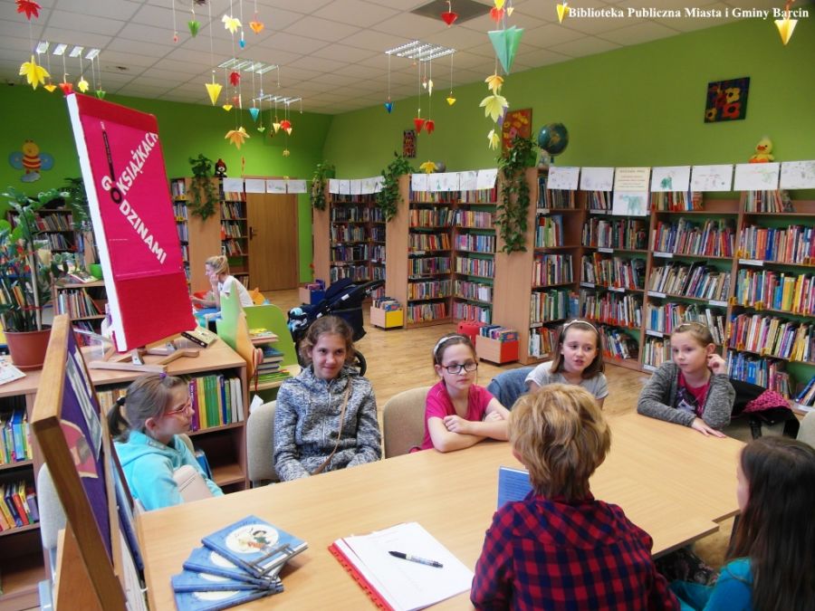 6 dzieci razem z pracownicą biblioteki prowadzącą spotkanie siedzą przy stole, i rozmawiają między sobą.