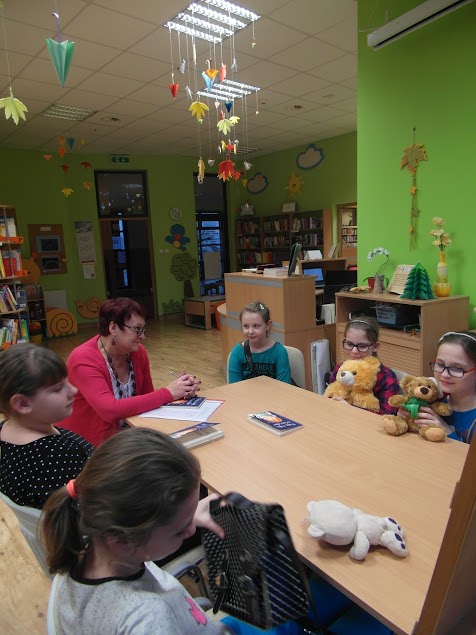 5 dziewczynek razem z bibliotekarką prowadzącą spotkanie siedzą wokół stołu, dwie dziewczynki trzymają w rękach pluszowe misie, inny miś leży na stole