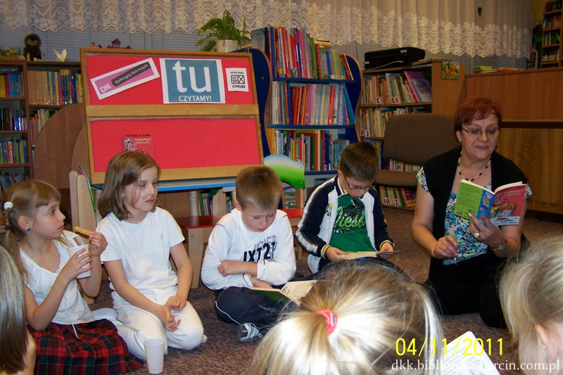 Grupa dzieci siedzi na dywanie, prowadząca spotkanie siedzi razem z nimi i czyta im ksiażkę