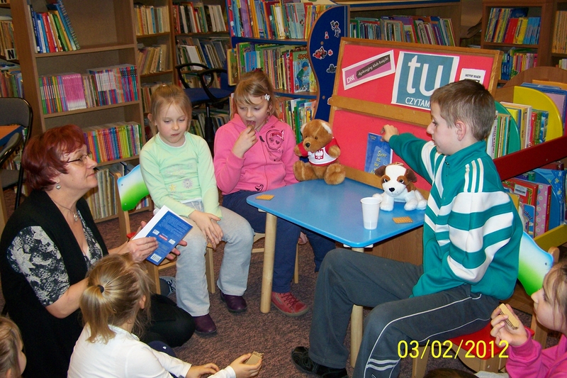 6 dzieci razem z bibliotekarką siedzą na krzesełkach, dzieci słuchają czytanej przez bibliotekarkę książki