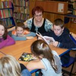 5 dzieci siedzi przy stole, jedno z dzieci czyta książkę, bibliotekarka prowadząca zajęcia przygląda się grupie.