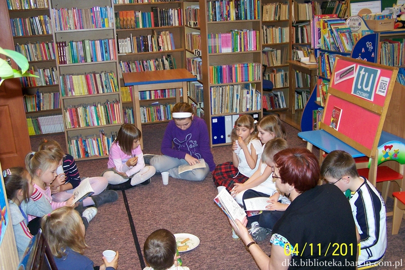 Grupa dzieci siedzi na dywanie, prowadząca spotkanie siedzi razem z nimi i czyta im ksiażkę