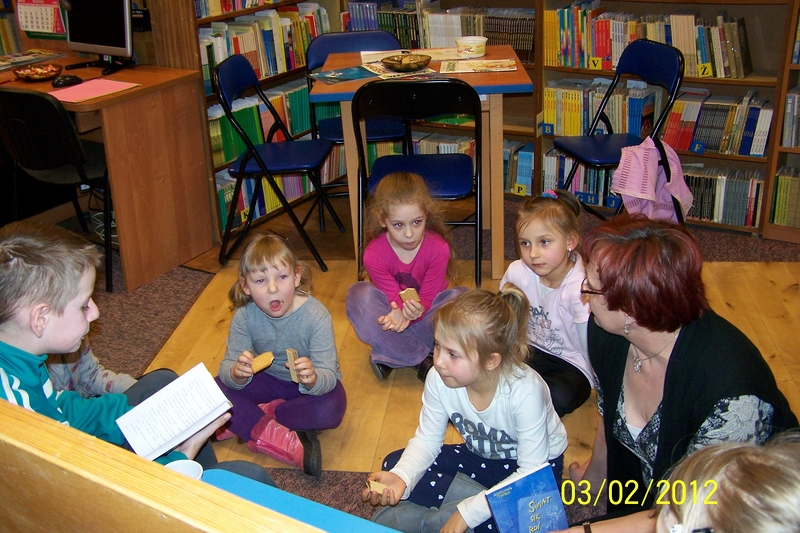 5 dzieci i bibliotekarka siedzą na podłodze, chłopiec czyta ksiażkę.