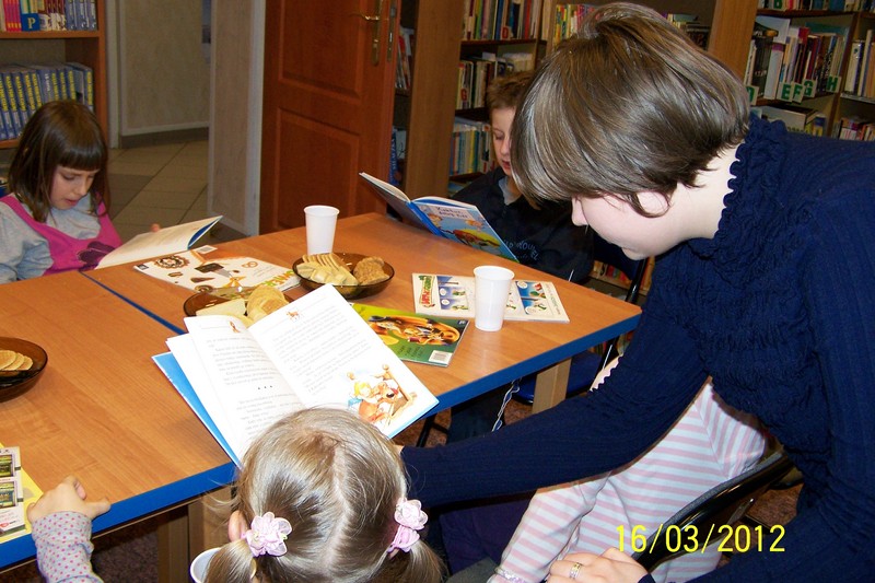 Dzieci siedzące przy stołach czytają książki, przy jednym z nich pochyla się dziewczyna ubrana w granatowy sweter.