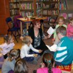 6 dzieci razem z bibliotekarką siedzą na krzesełkach, dzieci słuchają czytanej przez bibliotekarkę książki