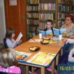 6 dzieci i bibliotekarka prowadząca spotkanie siedzą przy stołach, 3 dzieci i bibliotekarka czytają książki trzymane w rękach, inne dzieci słuchają, jedna dziewczynka pije sok z jednorazowego kubka