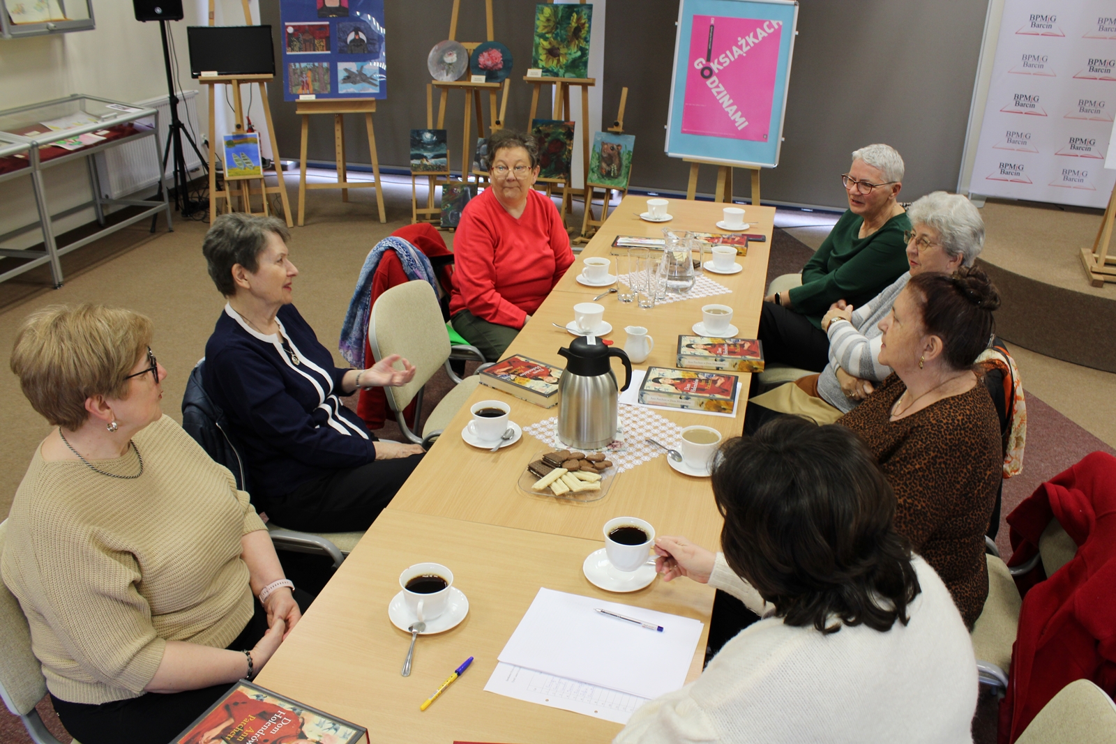 Grupa 7 kobiet w trakcie dyskusji na temat książki, siedzą przy stołach, popijają kawę i herbatę.