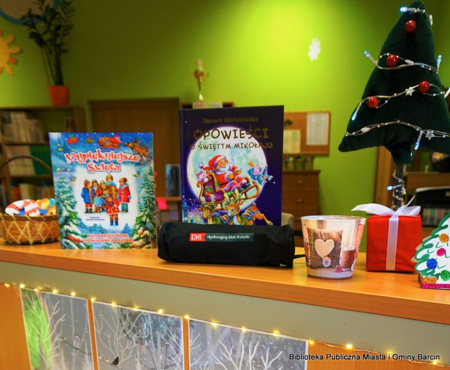 Książki o tematyce Świąt Bożego Narodzenia stojące na ladzie bibliotecznej, wokół książek stoją świąteczne osdoby
