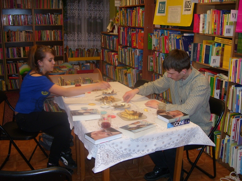dwójka nastolatków siedzi przy stole na którym leżą książki, stoją talerzyki z ciastkami i szklanki do napojów.