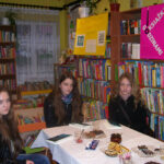 3 dziewczyny siedzące przy stole patrzą w stronę aparatu, na stole leżą książki, talerzyk z ciastkami, szklanki do herbaty