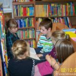 4 dzieci siedzi na ławce w bibliotece, chłopiec trzyma ksiażkę, dziewczynki przyglądają się tej ksiażce.