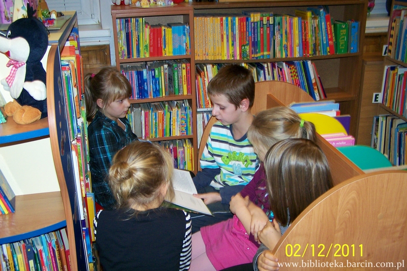 4 dzieci siedzi na ławce w bibliotece, chłopiec trzyma ksiażkę, dziewczynki przyglądają się tej ksiażce.