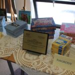 Stół z nagrodami dla uczestników konkursu, widać pamiątkowy grawerton dla zwycięskiej szkoły, gry planszowe, książki, pióra.