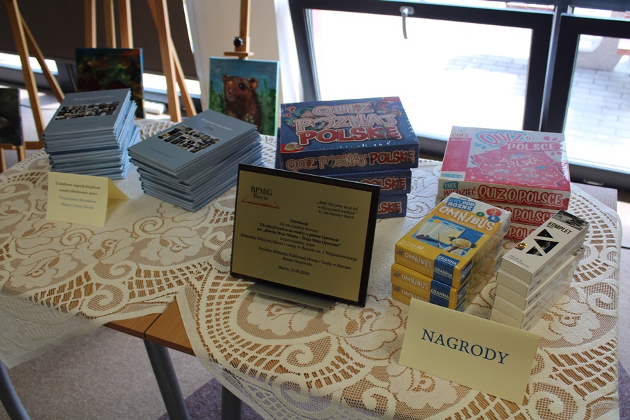 Stół z nagrodami dla uczestników konkursu, widać pamiątkowy grawerton dla zwycięskiej szkoły, gry planszowe, książki, pióra.