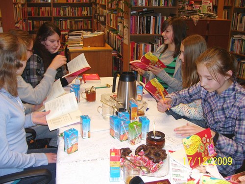 Grupa nastolatek siedzących przy stole z ksiażkami w ręku, na stole leżą ciastka, soki, termos z kawą lub herbatą.