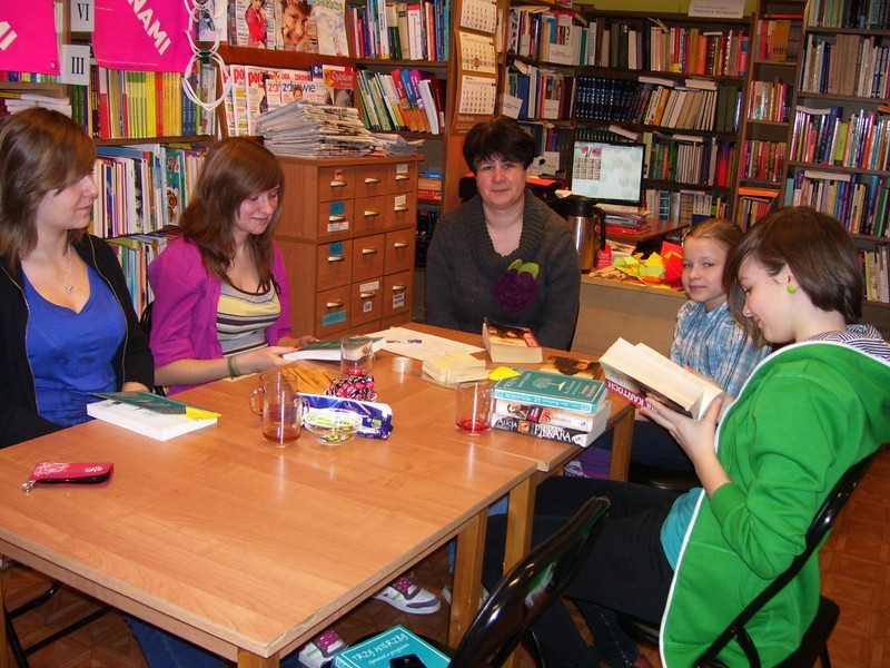 4 czytelniczki i bibliotekarka w czasie spotkania DKK, wszyscy siedzą przy stołach, dwie osoby mają w rękach ksiażkę