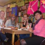 4 osoby, trzy nastolatki, jedna dorosła kobieta siedzą przy stole, patrzą w stronę aparatu, kobieta która jest koordynatorką DKK trzyma i pokazuje do aparatu okładkę ksiażki