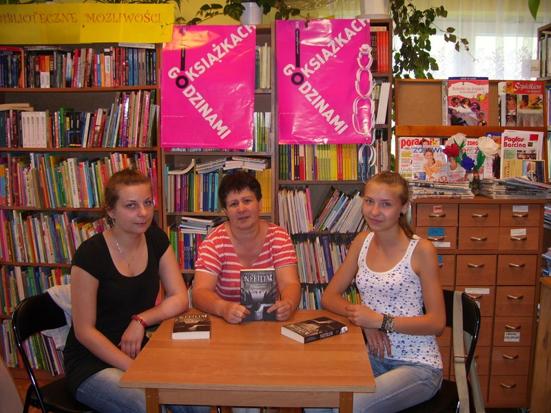 3 osoby przy stole, dwei nastolatki i prowadząca spotkanie bibliotekarka siedzą przy stole na którym leżą omawiane ksiażki, bibliotekarka pokazuje okładkę ksiażki do aparatu.