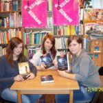 3 dziewczyny siedzące przy kwadratowym stoliku, w rękach trzymają przeczytane książki i pokazują ich okładki w stronę aparatu